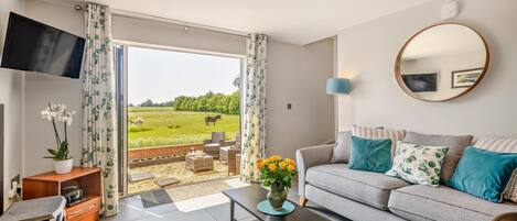 A modern living room with a grey sofa, a flat-screen TV, a coffee table, and a view of a green field with cows.
