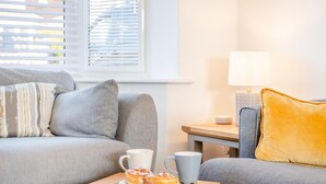 En hyggelig stue med en grå sofa, et lille træbord med et fad med kager og to kopper, en lampe og et vindue med persienner.