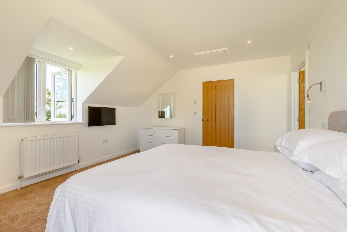 Ein Schlafzimmer mit einem großen Bett, einem an der Wand montierten Fernseher, einer Holztür und einem Fenster mit Vorhängen.