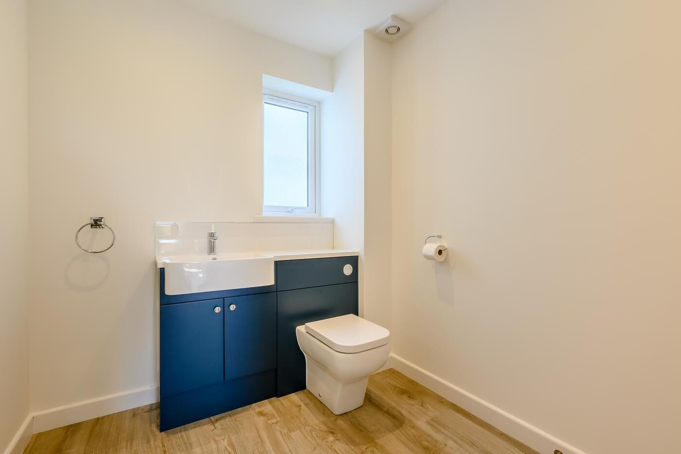 A small, modern bathroom with a white sink, blue cabinets, a white toilet, and a window above the sink.