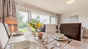 A dining area with a table set for four, including plates, bowls, and cutlery. There are two vases with flowers, a lamp, and a view of the outdoors through large windows. - 2 Bed in Saundersfoot (oc-s27727) (Saundersfoot)