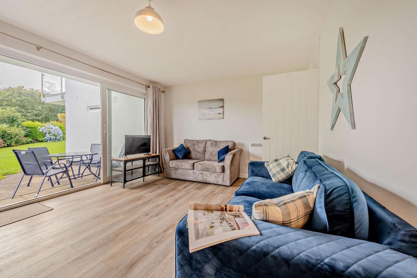Una sala de estar con un sofá azul, un sofá gris, un televisor sobre un mueble bajo y un comedor con sillas y una mesa.