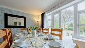 Un'area pranzo con un tavolo apparecchiato per quattro, sedie, un vaso con fiori e vista all'esterno attraverso ampie finestre.