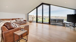 A spacious living room with a large brown leather sofa, a small coffee table, and a TV. The room has large glass doors leading to an outdoor area. - 4 Bed in Beadnell (oc-b27650) (Beadnell)