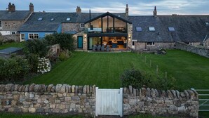 A modern house with a stone exterior, a well-maintained lawn, and a stone wall gate. - 4 Bed in Beadnell (oc-b27650) (Beadnell)