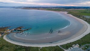 An aerial view of a coastal area with a sandy beach, a small harbor with boats, and surrounding green fields. - 4 Bed in Beadnell (oc-b27650) (Beadnell)