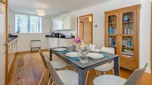 A modern kitchen with white cabinets, a dining table set for four, and a wooden bookcase filled with books. - 2 Bed in Southwold (oc-ftw2) (Southwold)