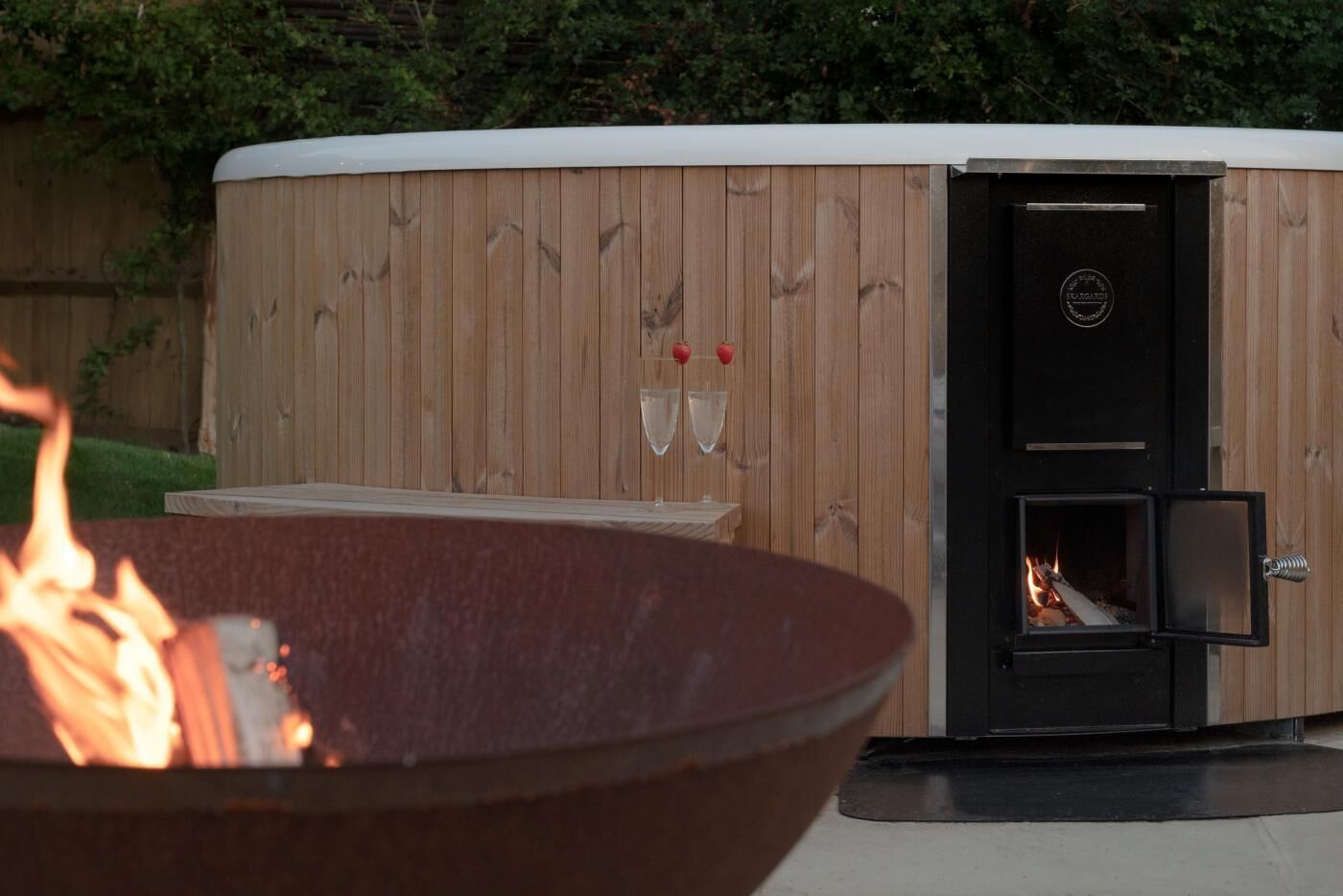 A wooden sauna with a glass door, a fire pit in the foreground, and a small table with two glasses and strawberries.
