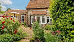 A stone house with a tiled roof, surrounded by lush greenery and flowering plants. - 3 Bed in Wiveton (oc-d19479) (Wiveton)
