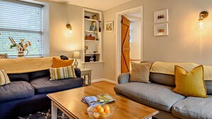 A cozy living room with two sofas, a coffee table with magazines and a bowl of fruit, a bookshelf, and a wooden door. - 3 Bed in Wiveton (oc-d19479) (Wiveton)