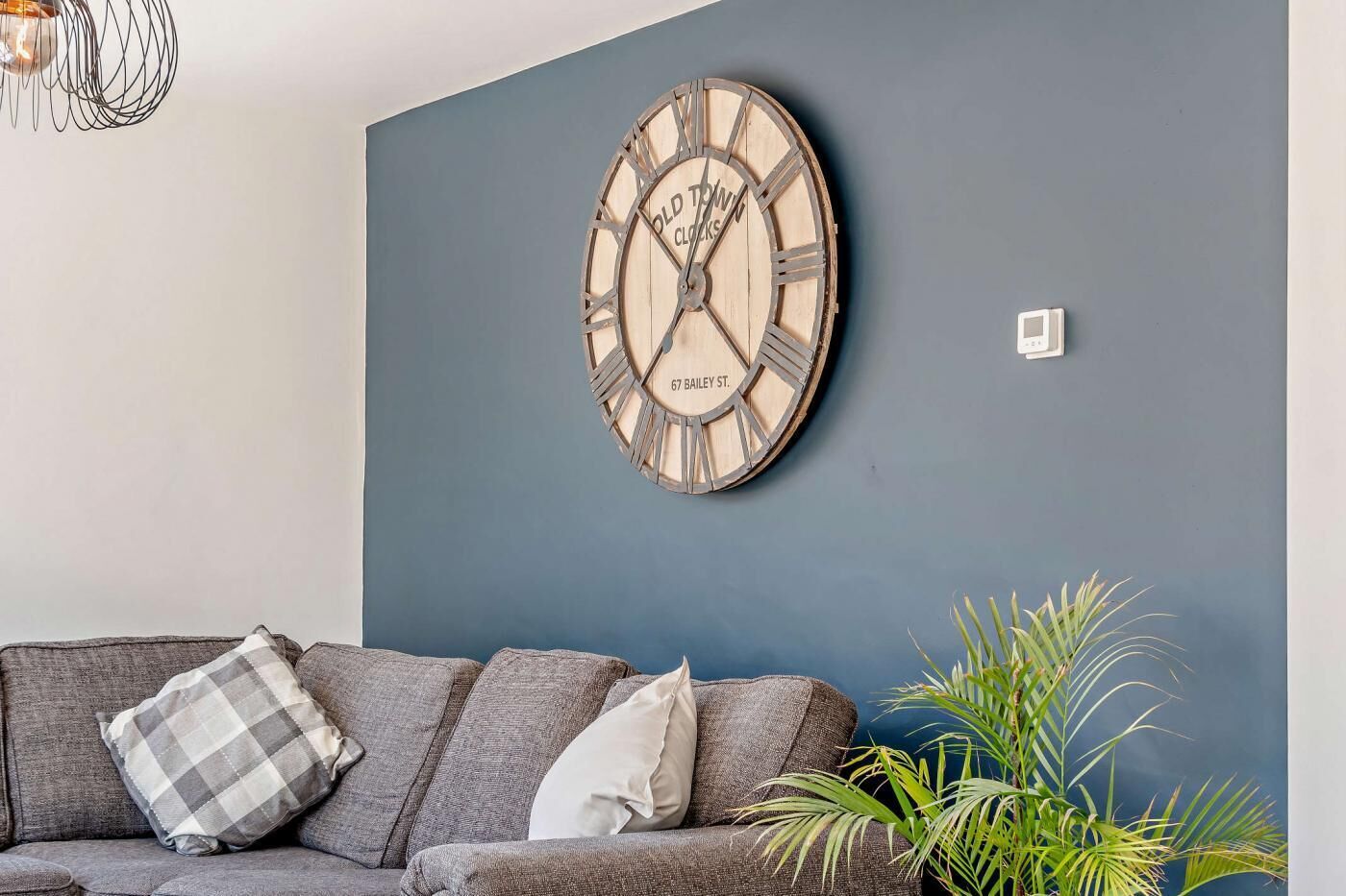 A living room with a large wooden clock on the wall, a grey sofa with cushions, and a potted plant.