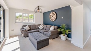 A modern living room with a sectional sofa, a grey ottoman, a wall clock, and a potted plant. - 3 Bed in Tholthorpe (oc-r23426) (Tholthorpe)