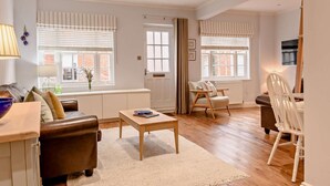 Ein geräumiges Wohnzimmer mit einer braunen Ledersofa, einem Holztisch und einem weißen Sessel. Zwei Fenster mit Jalousien, ein Heizkörper und ein Fernseher auf einem Schrank sind ebenfalls vorhanden.
