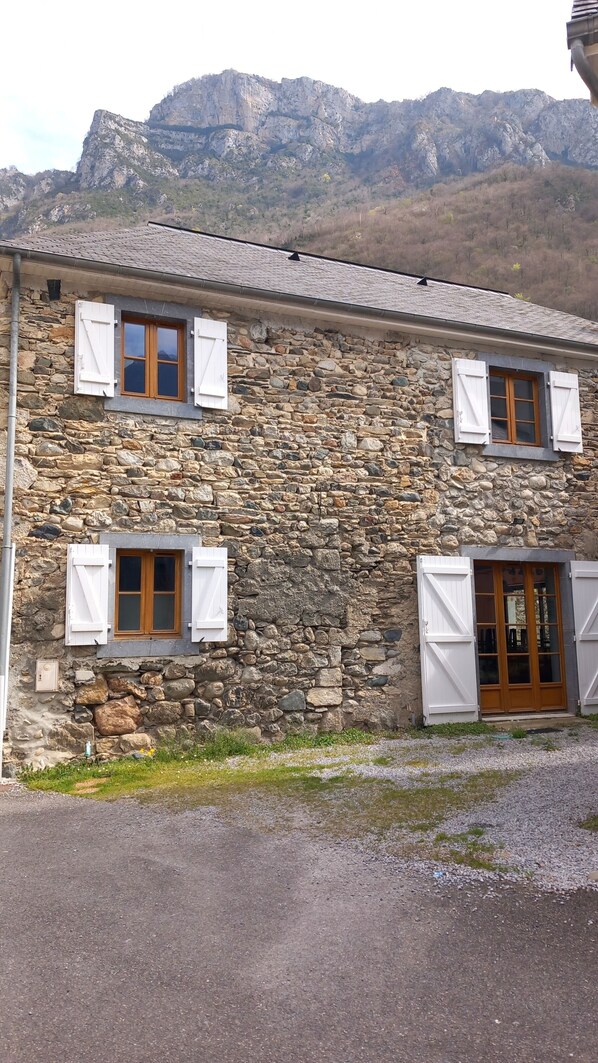 A stone building with two windows and a wooden door, set against a mountainous backdrop.