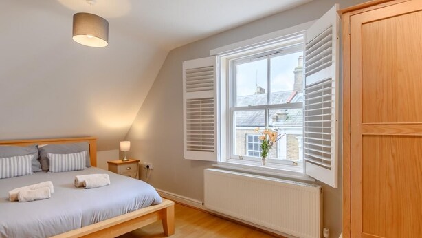 Een slaapkamer met een bed, nachtkastje, lamp, raam met luiken, radiator en een houten garderobe.