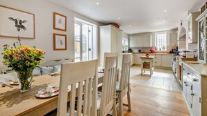 A modern kitchen with white cabinets, a wooden dining table, and chairs. There is a vase with flowers on the table. - 5 Bed in West Runton (oc-f27744) (West Runton)