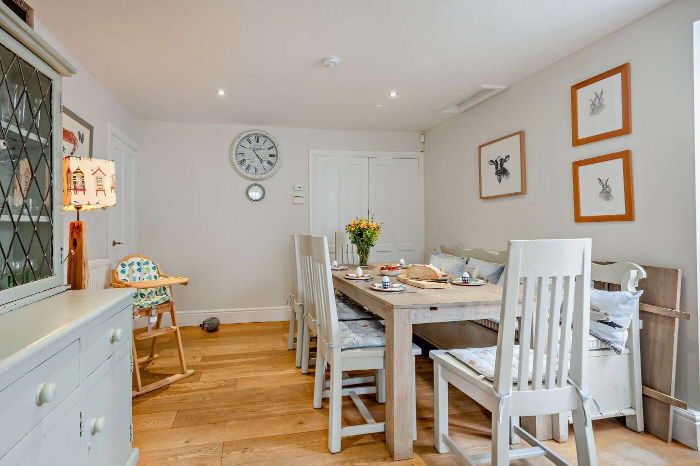 A dining area with a wooden table, white chairs, a clock on the wall, and framed pictures.