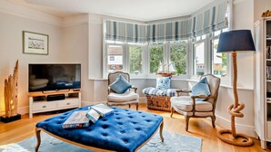A living room with a television, a blue ottoman, wicker chairs, and a large window with striped curtains. - 5 Bed in West Runton (oc-f27744) (West Runton)