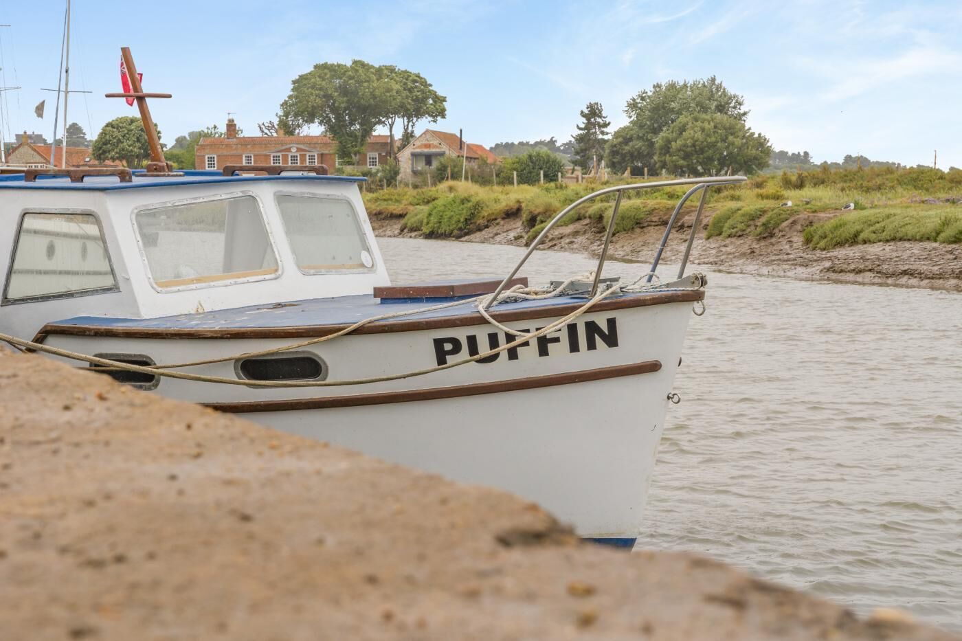 Un bateau nommé « PUFFIN » a accosté sur la rive d’une rivière, dont le rivage était sablonneux, avec des bâtiments visibles au loin.