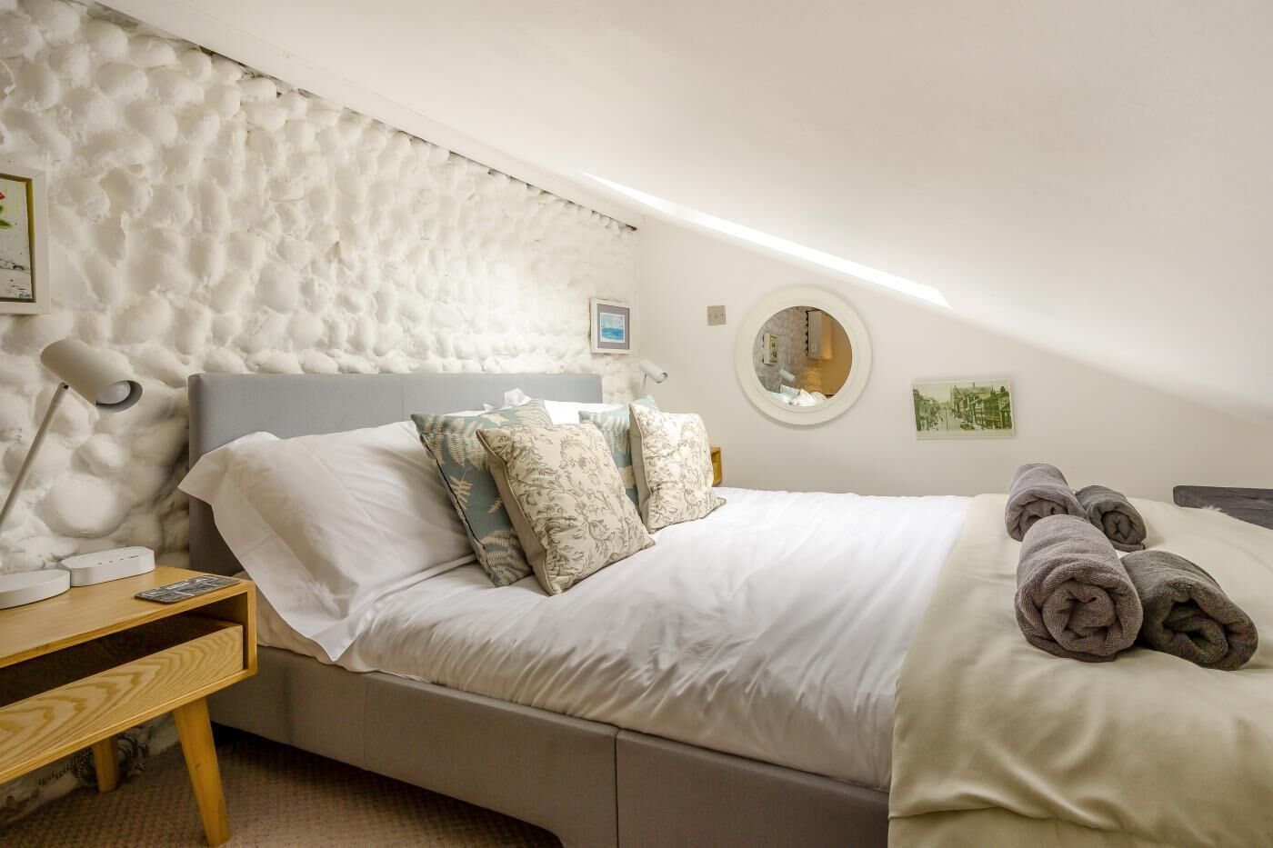 A bedroom with a bed, pillows, a bedside table with a lamp, a round mirror, and a wall-mounted picture.