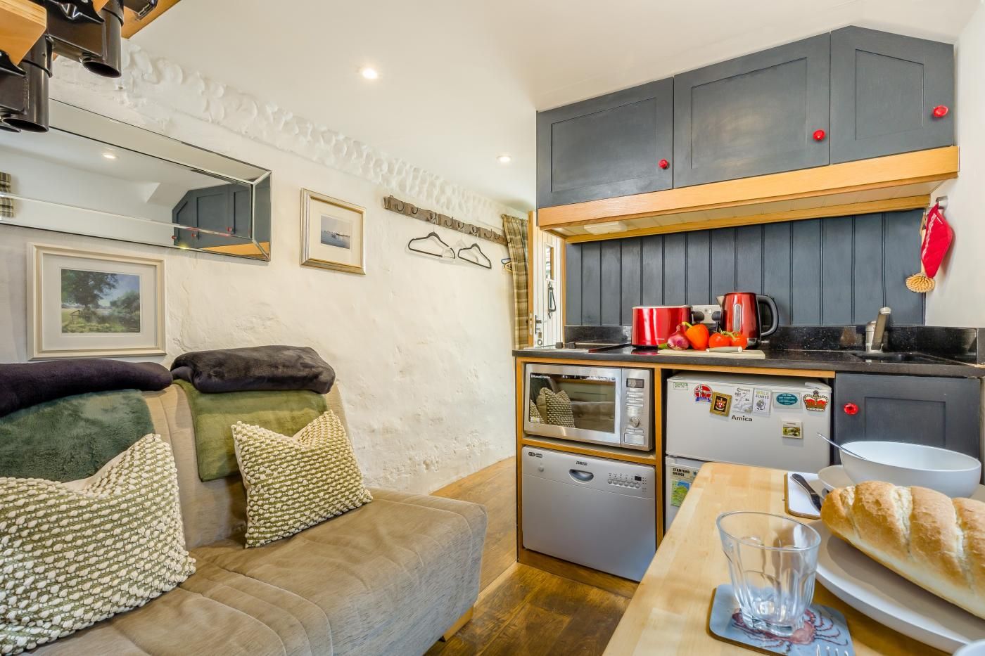 A compact living space with a kitchenette, a sofa with cushions, a dining table with a glass, and a red kettle.