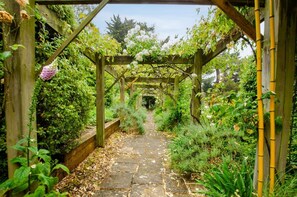 A stone pathway leading through a wooden pergola garden with lush greenery and flowering plants. - 4 Bed in Aldeburgh (oc-h27787) (Aldeburgh)