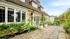 Ett traditionellt hus med en stenlagd uteplats, stora fönster och en trädgård med olika växter och blommor.