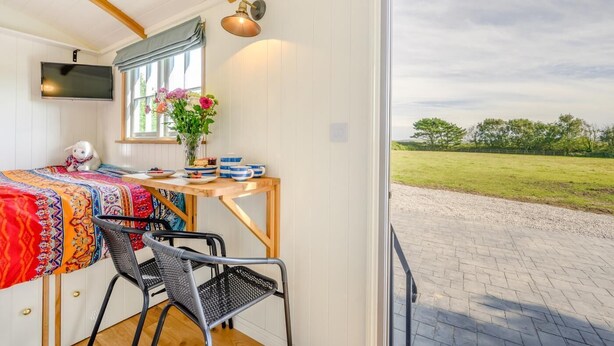 Ein gemütliches Zimmer mit einem Bett, einem Tisch mit Blumenvase, einem Sessel und einem an der Wand befestigten Fernseher.