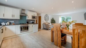 A modern kitchen with white cabinets, a built-in oven, and a microwave. There is a dining area with a wooden table and chairs, and a large window providing natural light. - 2 Bed in St. Issey (oc-o27778) (Saint Issey)