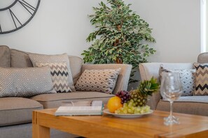 A living room with a sofa, patterned cushions, a wooden coffee table with a book and fruit, and a large potted plant.