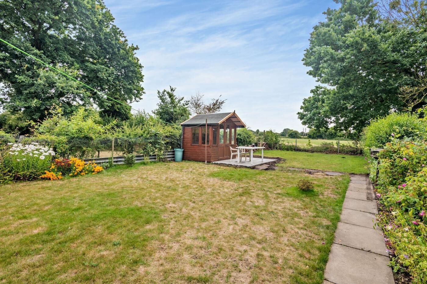 A small wooden structure with a roof, possibly a shed or gazebo, in a grassy backyard with a paved path and a variety of plants and flowers.