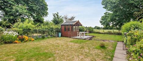 A small wooden structure with a roof, possibly a shed or gazebo, in a grassy backyard with a paved path and a variety of plants and flowers.