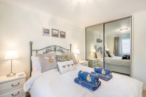 A neatly made bed with pillows, a bedside table with a lamp, a mirror, and a window with curtains.