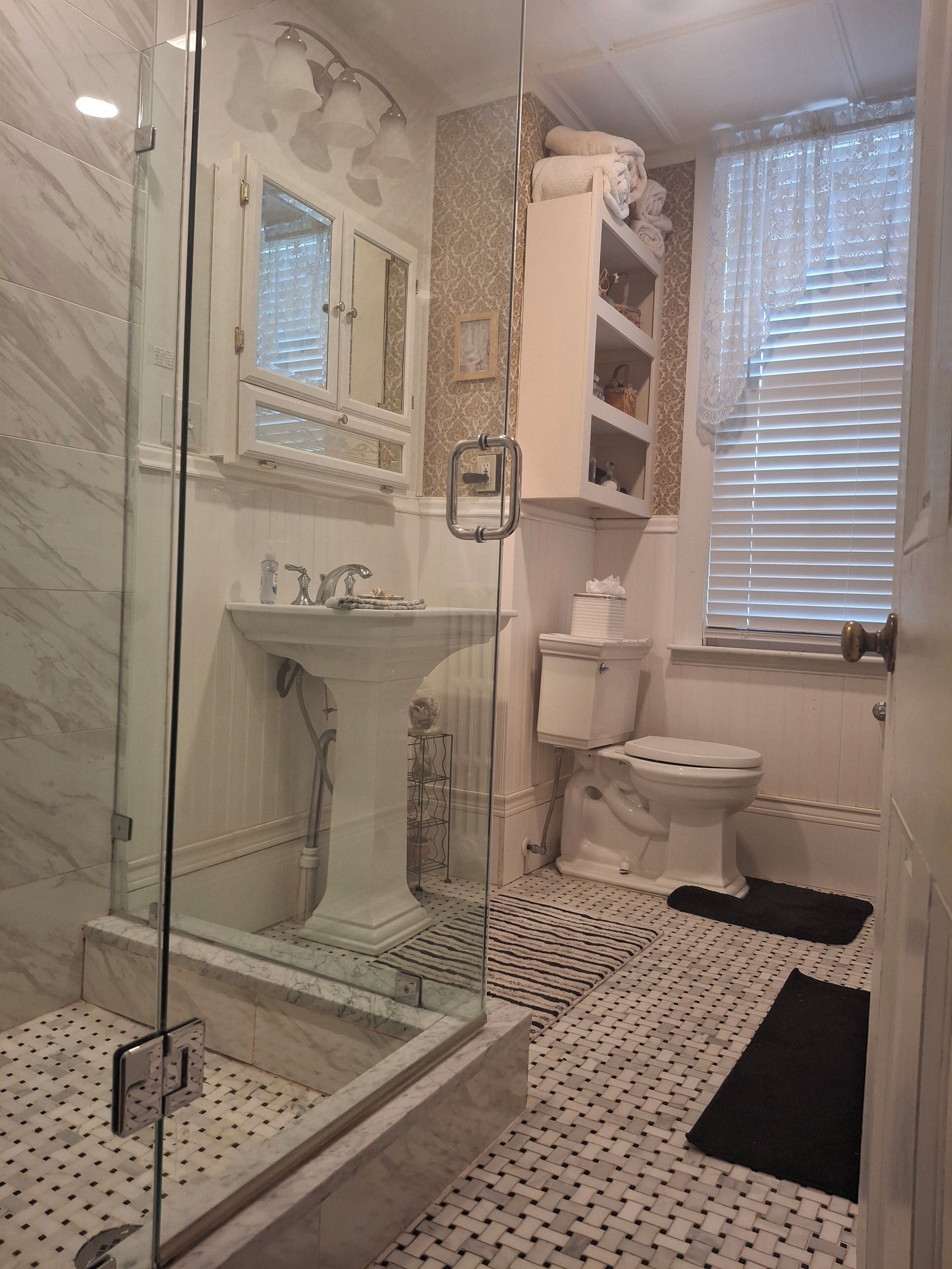 A bathroom with a glass-enclosed shower, a white sink, a toilet, and a window with blinds.