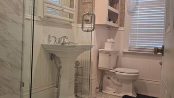 A bathroom with a glass-enclosed shower, a white sink, a toilet, and a window with blinds.