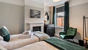 Une chambre d’hôtel avec deux lits, un fauteuil vert, une cheminée et une grande fenêtre avec des rideaux.