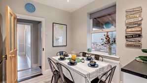 Un coin repas avec une table dressée pour quatre personnes, une horloge au mur et une fenêtre donnant sur un beau paysage.