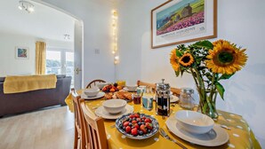 Un'area pranzo con un tavolo giallo apparecchiato per la colazione, con frutta, pasticcini e caffè. Si nota un quadro appeso alla parete e un vaso con girasoli.