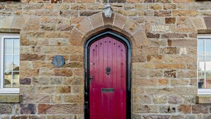 A red door with a black handle, set in a stone building with a window to the left. - 4 Bed in Broad Haven (oc-t27866) (Broad Haven)