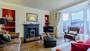 A living room with a fireplace, leather sofas, a television, and a large window with a view of parked cars. - 4 Bed in Broad Haven (oc-t27866) (Broad Haven)