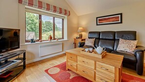 Ein gemütliches Wohnzimmer mit Fernseher, Couchtisch aus Holz, Ledersofa und einem großen Fenster mit Blick ins Grüne.
