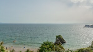 A coastal scene with a rocky outcrop in the water, a beach with people, and a pier with a building. - 2 Bed in Tenby (oc-s27901) (Tenby)