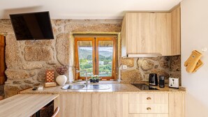 A kitchen with a stone wall, wooden cabinets, a TV mounted above the counter, a window with a view of greenery, and a sink.