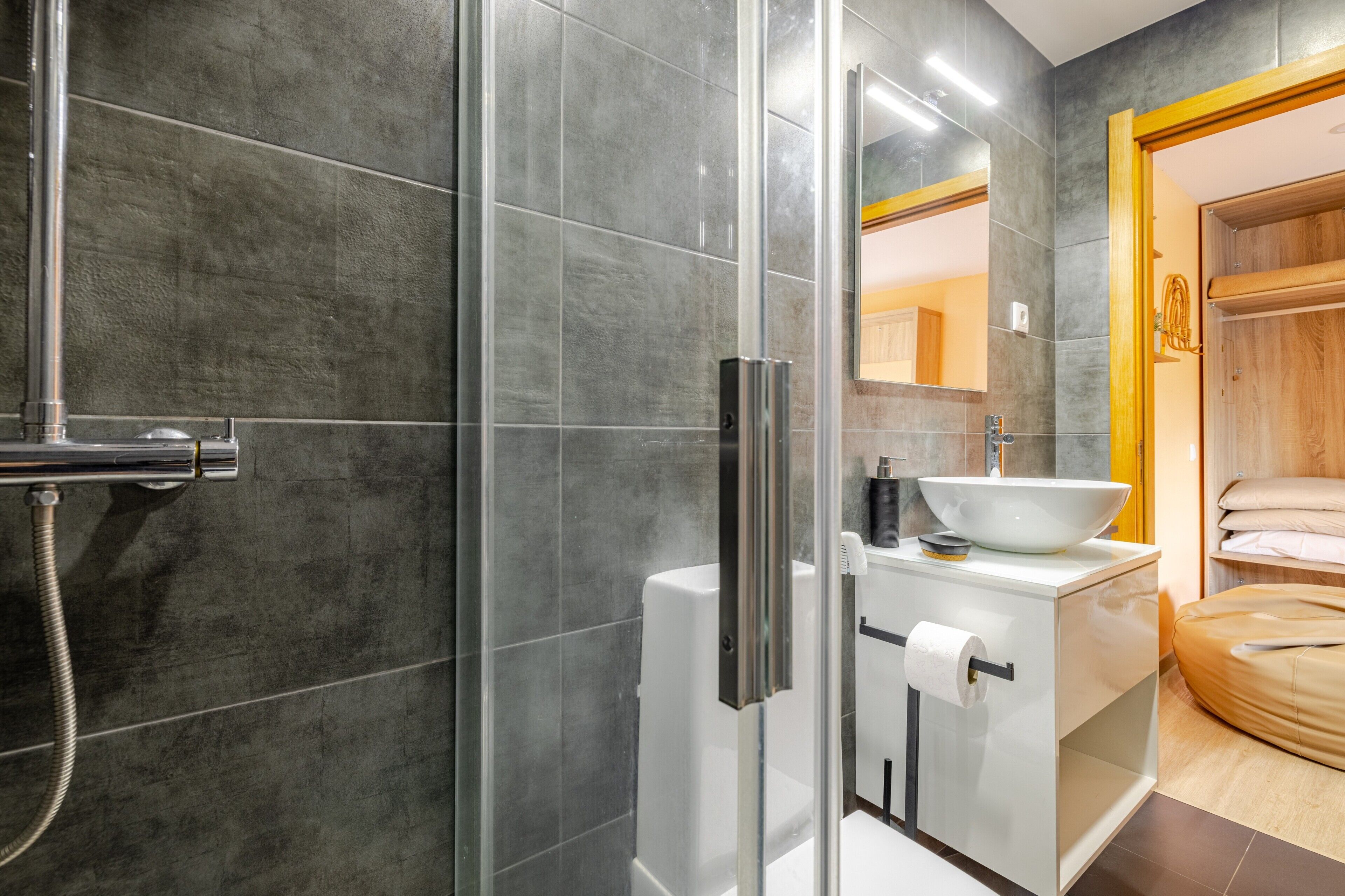 Un baño moderno con cabina de ducha de vidrio, un lavabo blanco con cuenco y una puerta de madera.