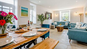 Amplio salón con mesa de comedor lista para una comida, un sofá azul y un ventanal que proporciona luz natural.