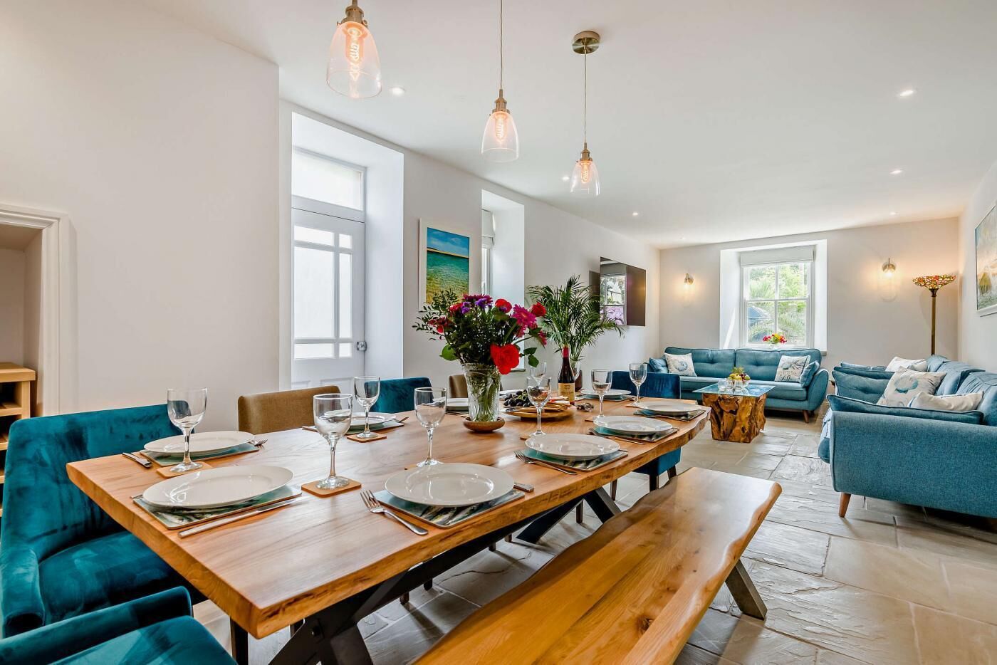 Een eethoek met een houten tafel gedekt voor vier personen, omringd door teal bekleden stoelen. Boven de tafel hangen hanglampen, en op de tafel staat een vaas met bloemen.