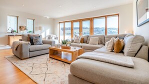 A spacious living room with a large sofa set, a coffee table, and a view of the city through large windows. - 4 Bed in Newquay (oc-b27906) (Newquay)