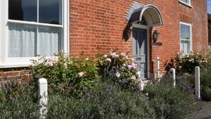 Ein rotes Backhaus mit weißer Tür, einer kleinen Veranda und einem Garten mit Lavendel und Rosen.