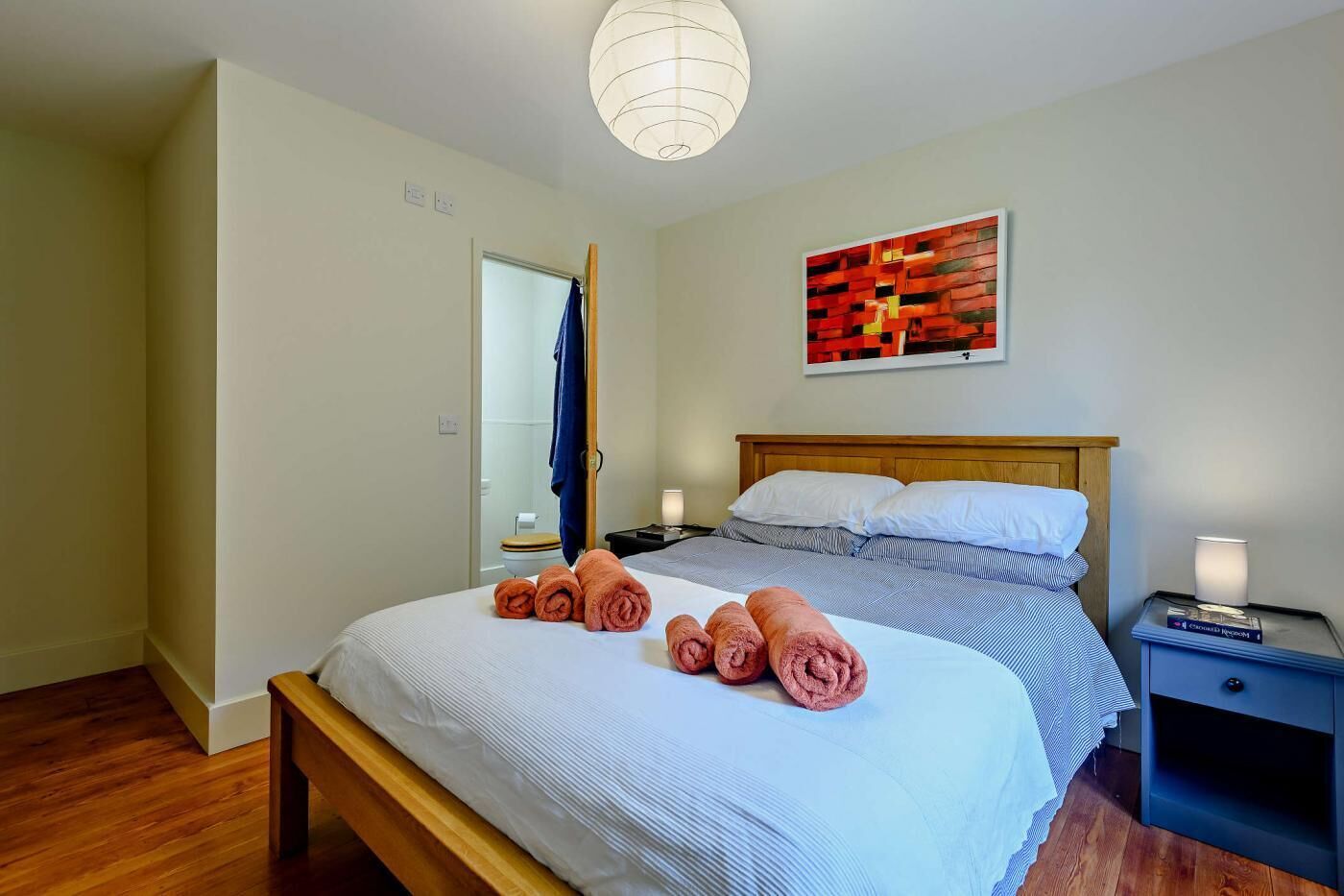 Ein Schlafzimmer mit Bett, Nachttisch, Nachttischlampe, einem Wandbild und einer Tür zu einem Badezimmer.