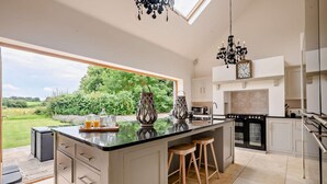 A modern kitchen with a large island, two bar stools, a chandelier, and a view of the garden. - 5 Bed in Ellough (oc-d8656) (Ellough)